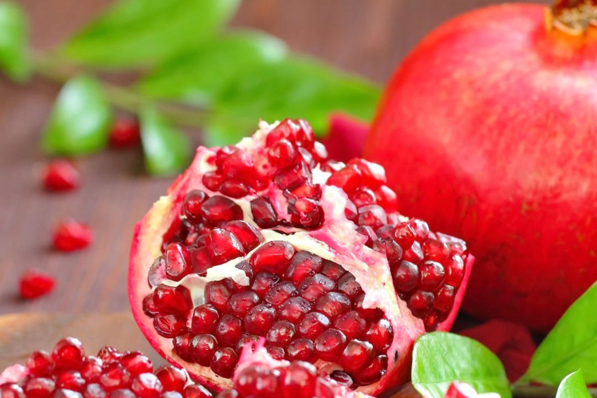 Two pomegranates one of which is cut open with seeds scattered on surface with green leaves.