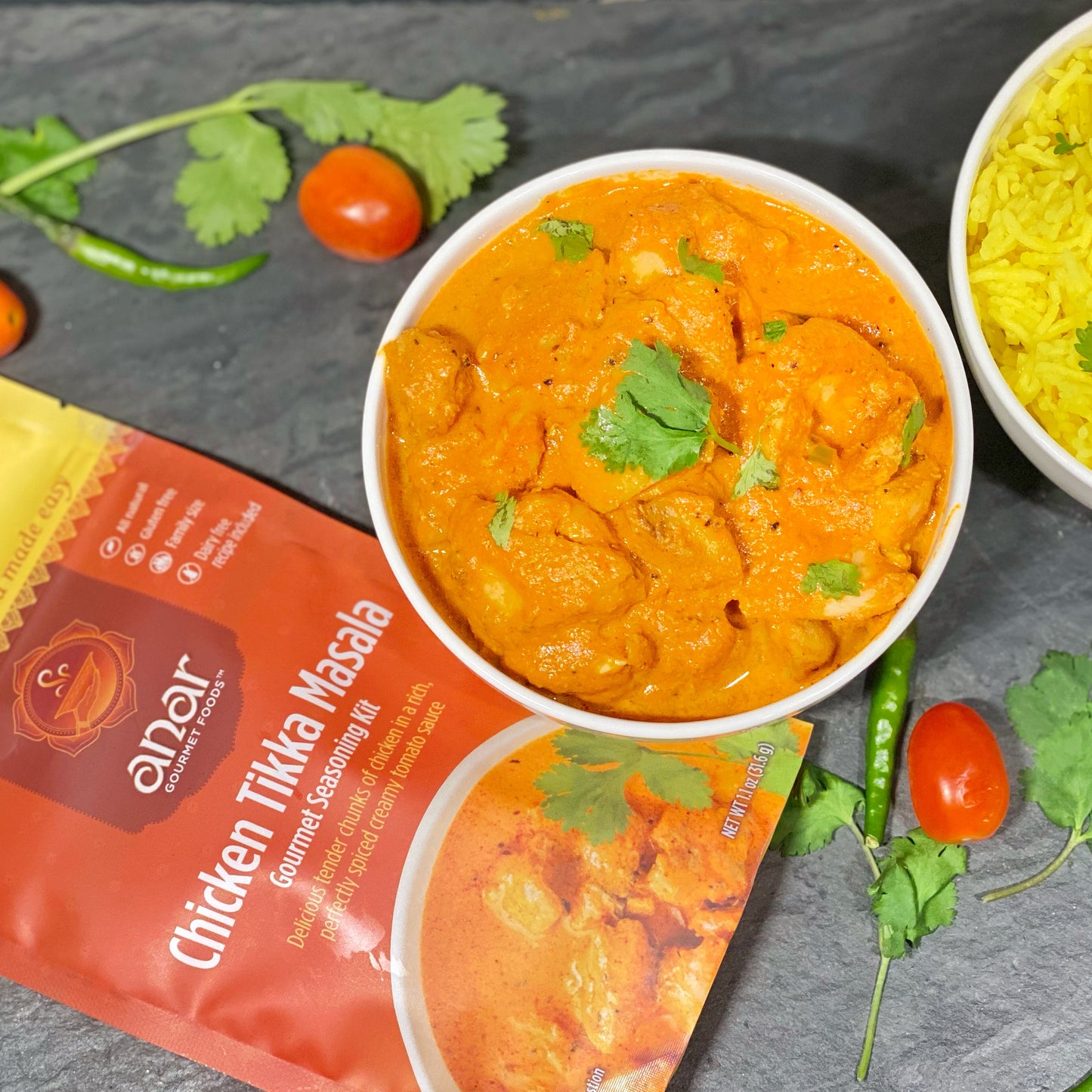 A packaged all natural, gluten free gourmet seasonings packet labeled 'Chicken Tikka Masala' alongside a prepared dish in a bowl, garnished with cilantro, with tomatoes and spices in the background.