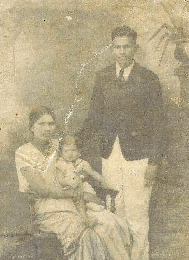 Vintage sepia-toned family portrait showing a seated woman holding a child next to a standing man in formal attire, with a mottled studio backdrop and visible age-related wear and creases.