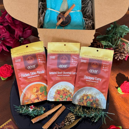Anar Gourmet Foods Indian seasoning blends gift box with holiday styling — Chicken Tikka Masala, Ground Beef Keema Curry, and Chickpea Curry packets with pinecones and poinsettia